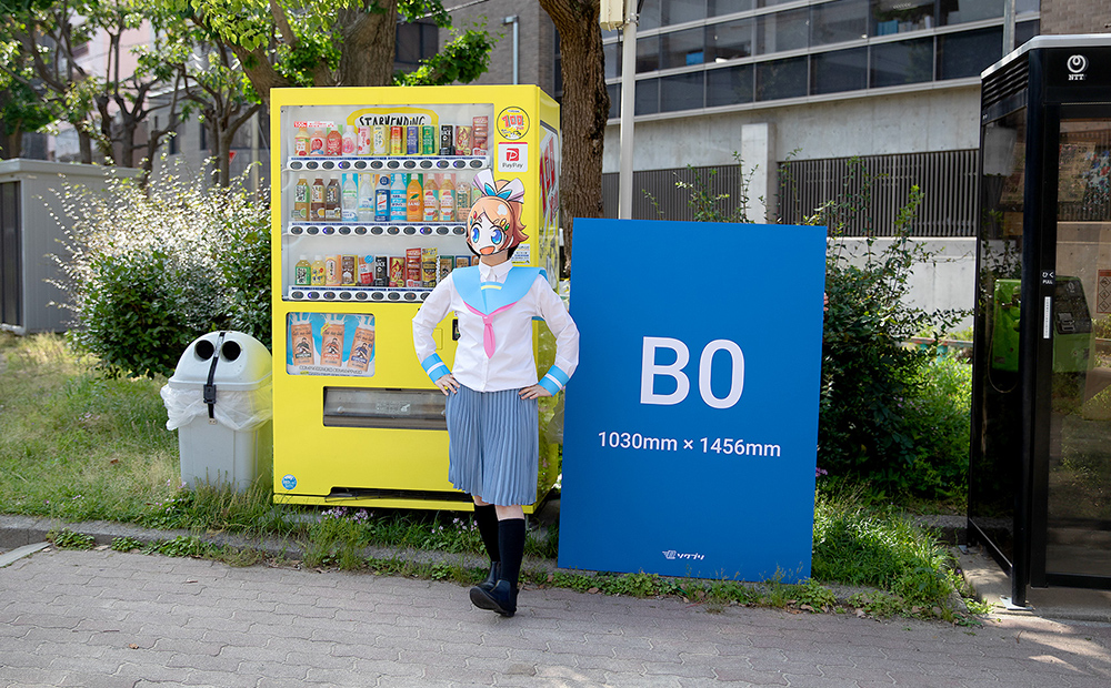 自動販売機とB0サイズの発泡パネルを並べた前に立っているソクプリ子ちゃん