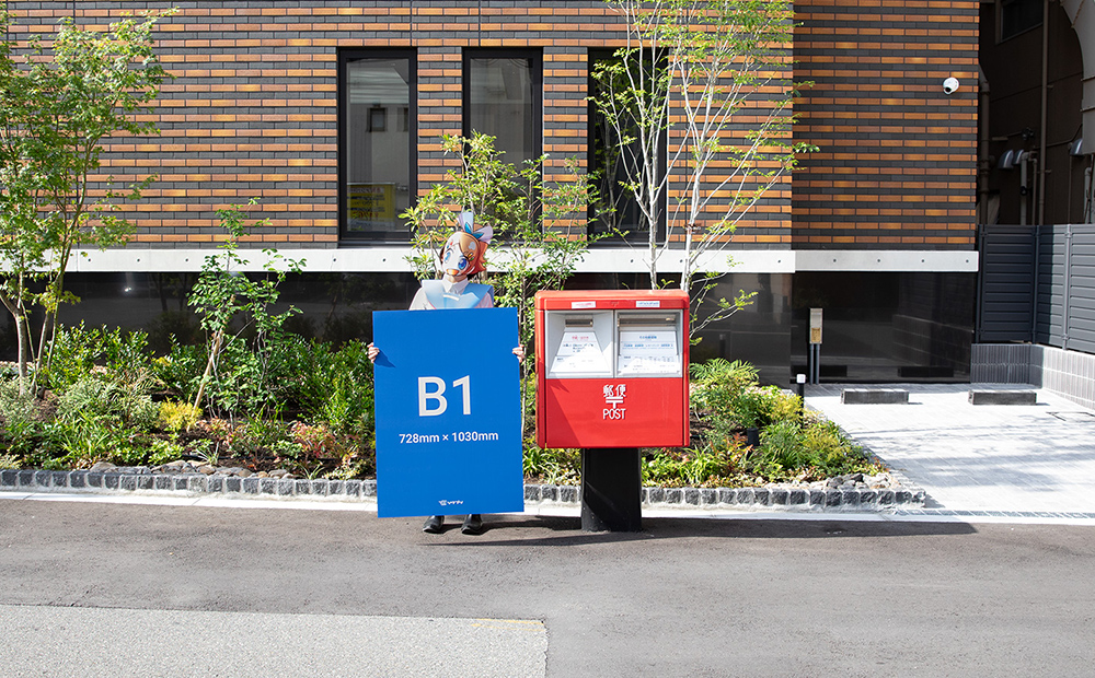 B1サイズの発泡パネルと二口のポストのサイズを比べているソクプリ子ちゃん