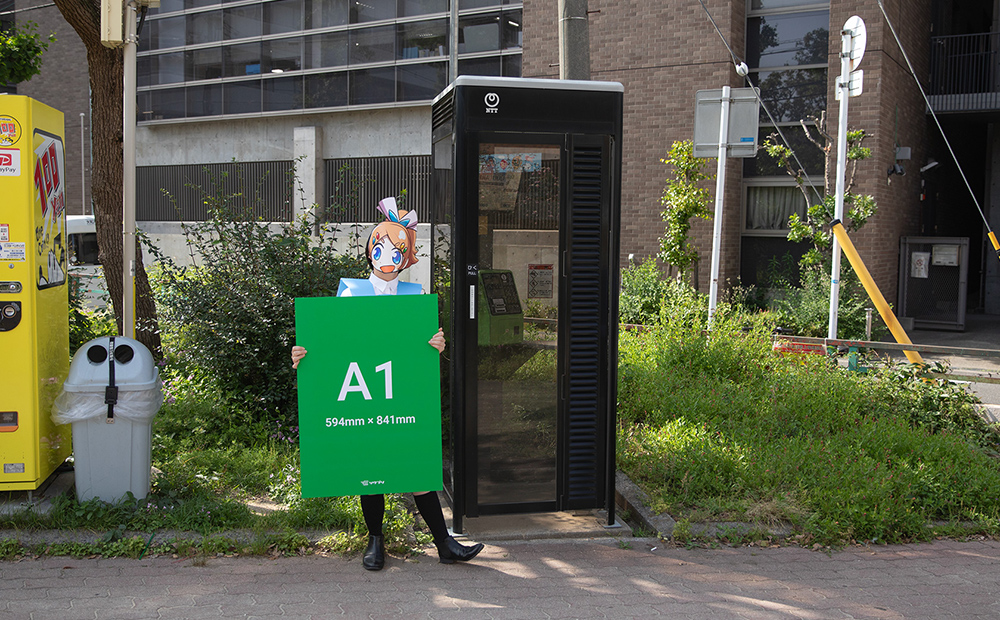 A1サイズの発泡パネルを手にして、電話ボックスの横に立ちサイズを比べているソクプリ子ちゃん