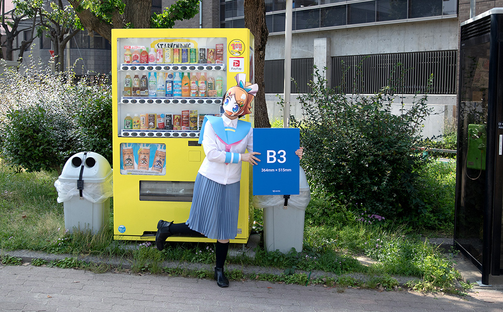 自動販売機の前でB3サイズの発泡パネルを手にして、サイズを比べているソクプリ子ちゃん